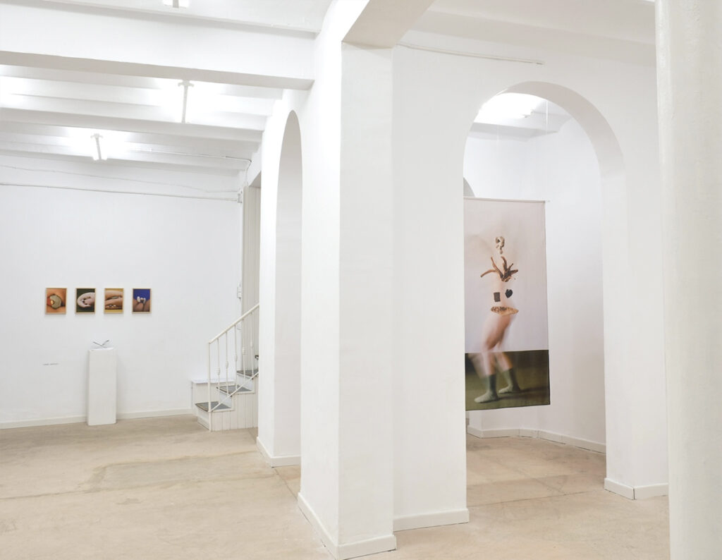 Vista general de una sala de Sorondo Projects con fotografías, una instalación sobre pedestal y una obra textil suspendida entre arcos blancos.