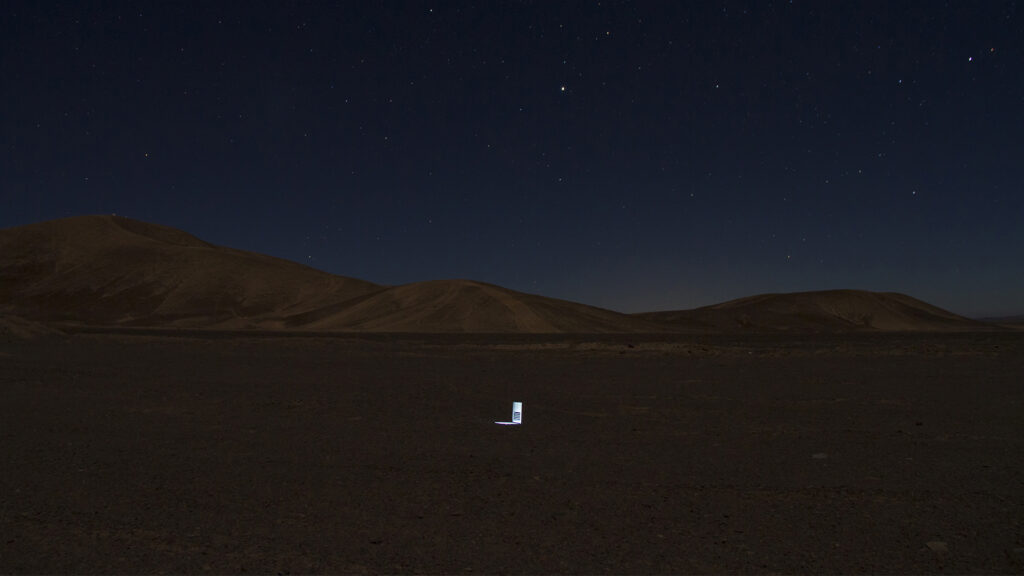 A nighttime desert landscape under a starry sky, with a small, illuminated object in the center of the scene.