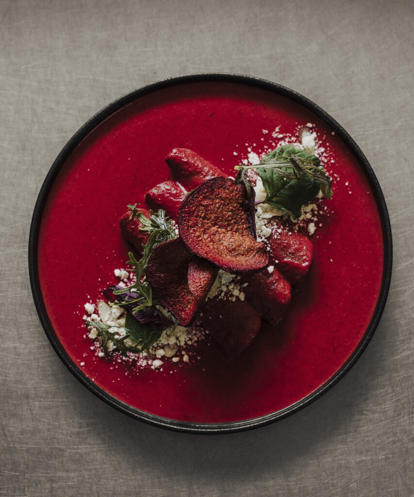 Platillo de mole rosa presentado en un plato negro, acompañado de betabel, queso, hojas frescas y otros elementos que contrastan con el intenso color de la preparación.