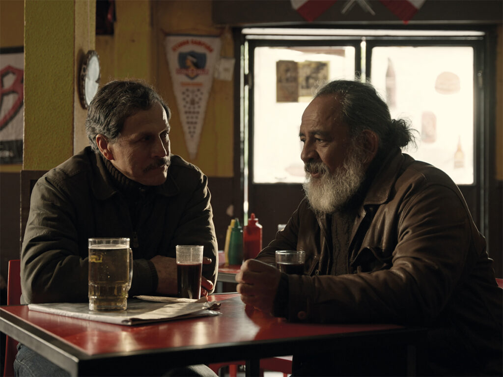 Dos hombres conversan sentados frente a una mesa en un bar, con vasos y botellas sobre la mesa.