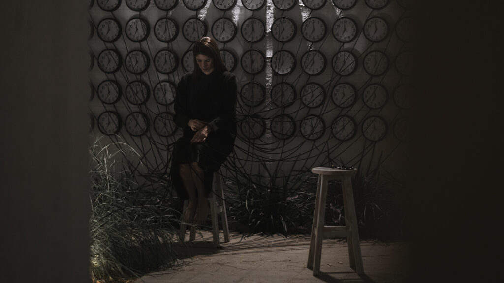 Mariajosé Fernández-Plenge durante una performance frente a una instalación compuesta por relojes conectados mediante cables, en un espacio expositivo.