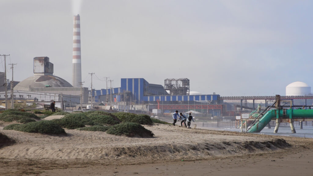 Paisaje costero de Loncura con jóvenes caminando por la playa y grandes instalaciones industriales y chimeneas al fondo.