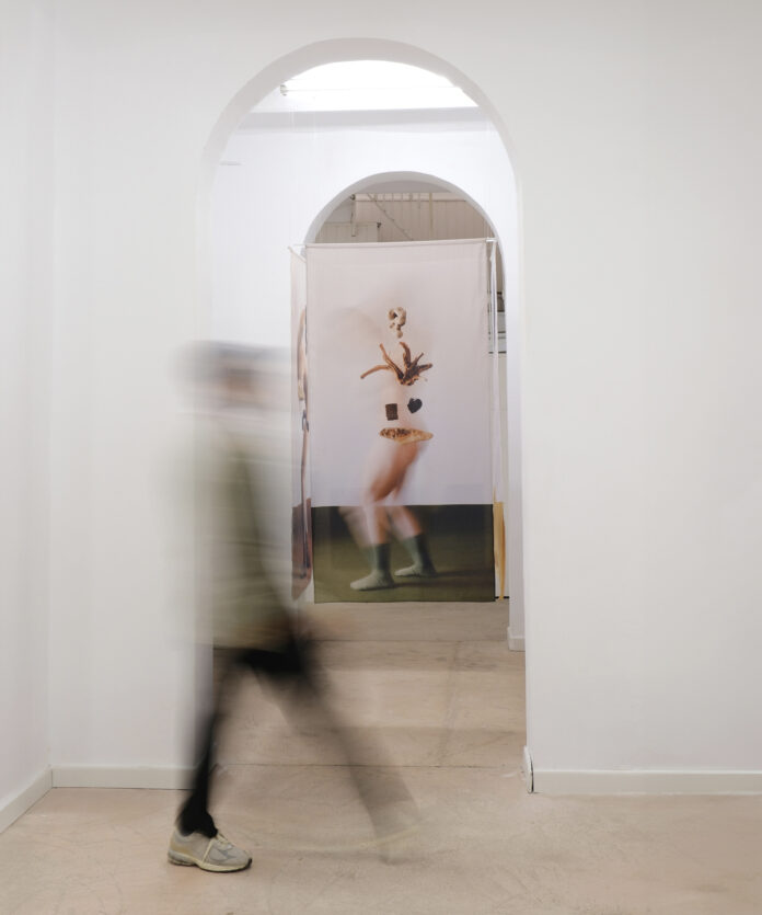 Persona captada en movimiento mientras atraviesa una sala de Sorondo Projects; al fondo se observa una obra textil suspendida enmarcada por un arco blanco.