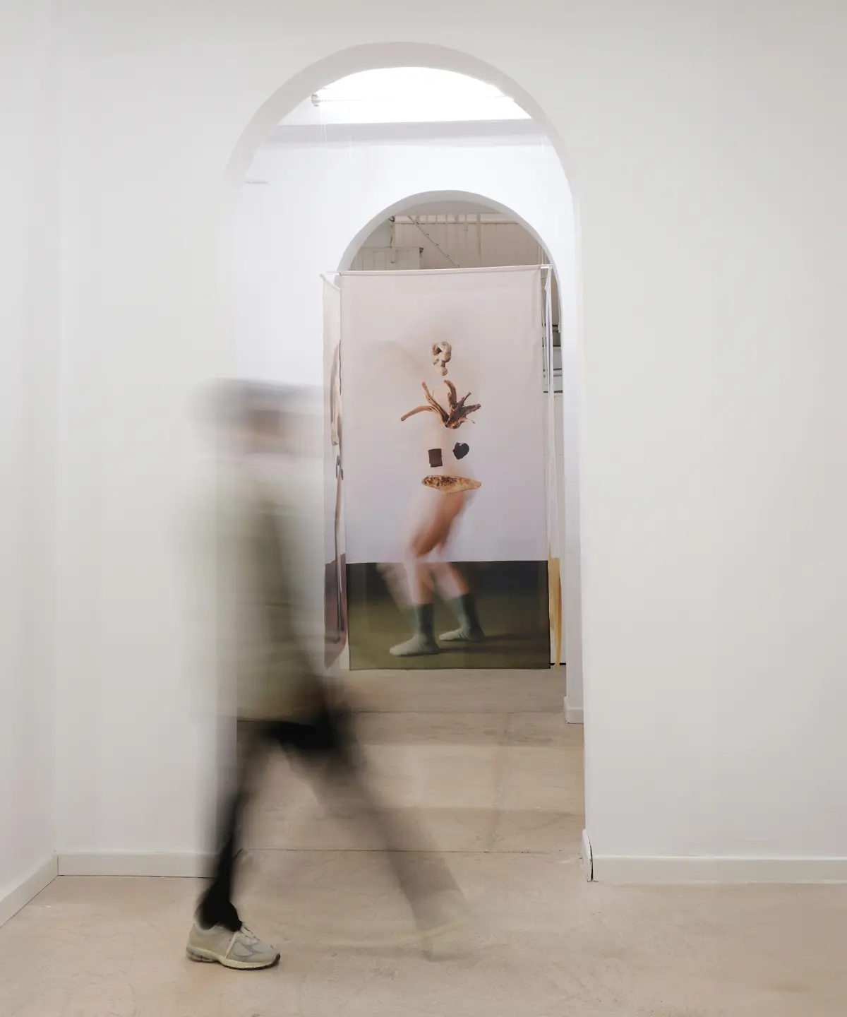 Persona captada en movimiento mientras atraviesa una sala de Sorondo Projects; al fondo se observa una obra textil suspendida enmarcada por un arco blanco.