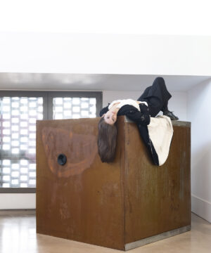 Mariajosé Fernández-Plenge reclining on a cubic wooden structure as part of an art installation in an exhibition space.