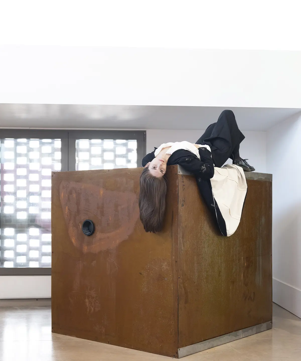 Mariajosé Fernández-Plenge recostada sobre una estructura cúbica de madera como parte de una instalación artística en un espacio expositivo.