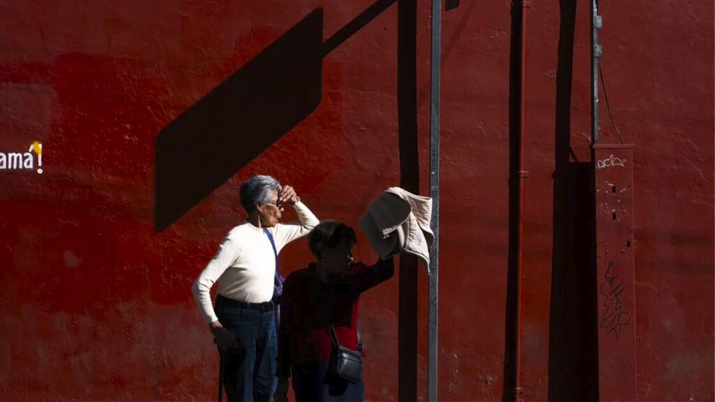 Fotografía de Gregorio Díaz de dos personas frente a un muro rojo intenso, atravesado por grandes sombras geométricas; una de ellas se cubre del sol con la mano y la otra sostiene una prenda sobre su cabeza.