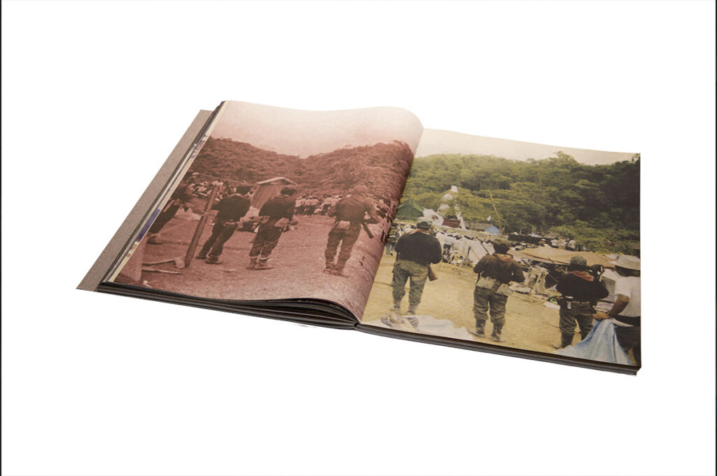 Fotolibro abierto sobre fondo blanco, con una fotografía a doble página de varias personas caminando entre un campamento rodeado de vegetación; una página está impresa en tonos rojizos y la otra a color.