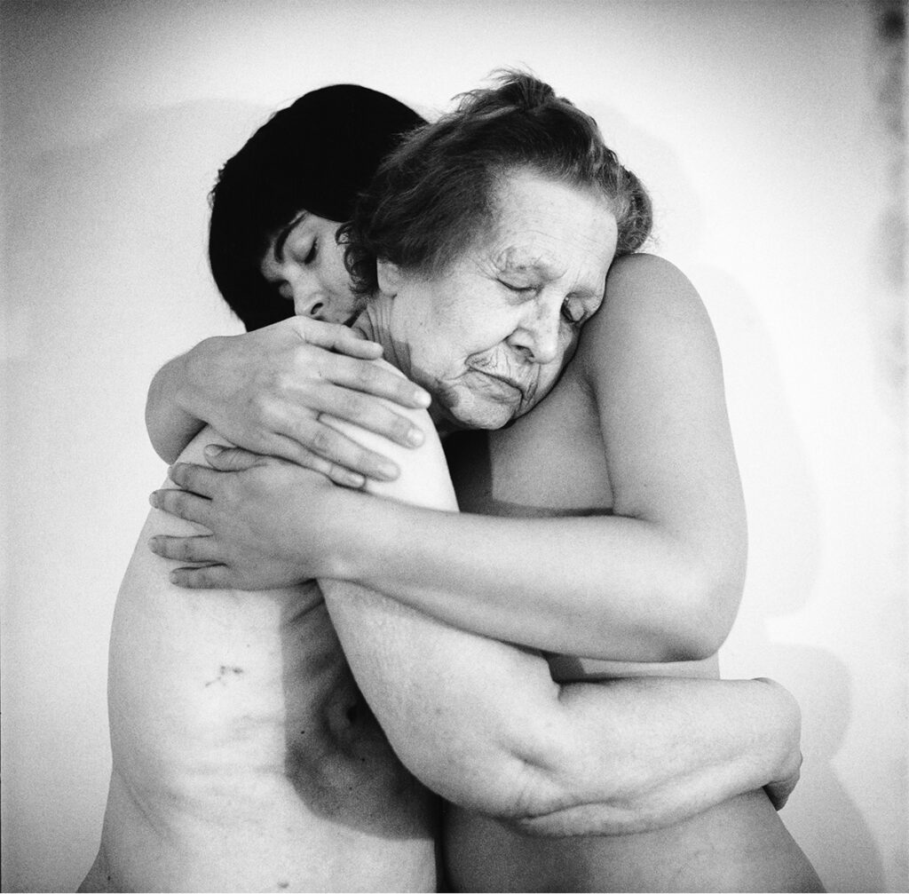 Fotografía en blanco y negro de dos personas desnudas abrazadas; una mujer mayor descansa el rostro sobre el hombro de la otra persona.