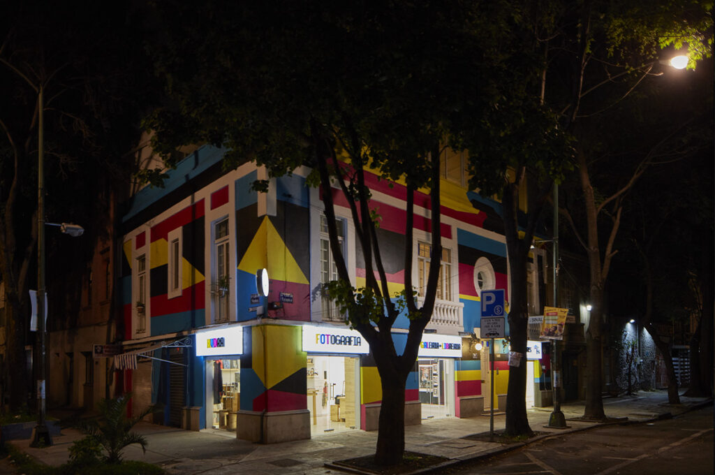 Fachada nocturna de Hydra Editorial en la Ciudad de México, con muros de colores y árboles frente al edificio iluminado.