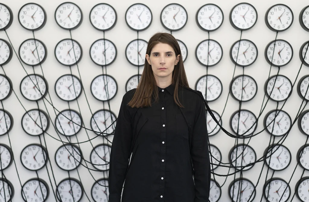 Retrato de Mariajosé Fernández-Plenge frente a una instalación formada por múltiples relojes conectados entre sí.