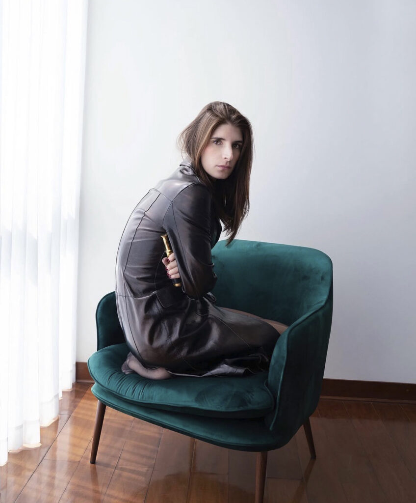Retrato de la artista Mariajosé Fernández-Plenge sentada en un sillón verde dentro de un espacio iluminado por luz natural.