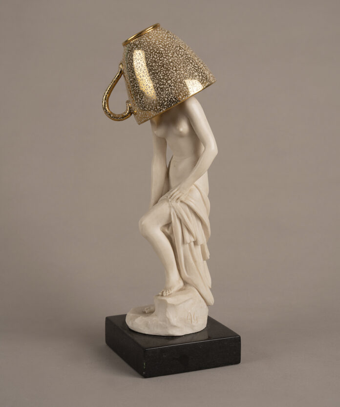 Figura escultórica femenina sobre una base oscura, intervenida con una taza de acabado dorado que cubre la cabeza. La pieza forma parte de Escenarios domésticos, proyecto en el que Marianna Dellekamp transforma objetos provenientes de su archivo familiar para explorar la memoria y los vínculos afectivos con las pertenencias.