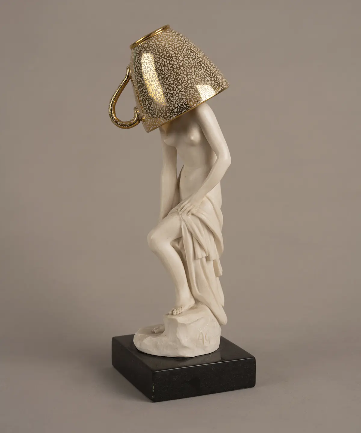 Figura escultórica femenina sobre una base oscura, intervenida con una taza de acabado dorado que cubre la cabeza. La pieza forma parte de Escenarios domésticos, proyecto en el que Marianna Dellekamp transforma objetos provenientes de su archivo familiar para explorar la memoria y los vínculos afectivos con las pertenencias.