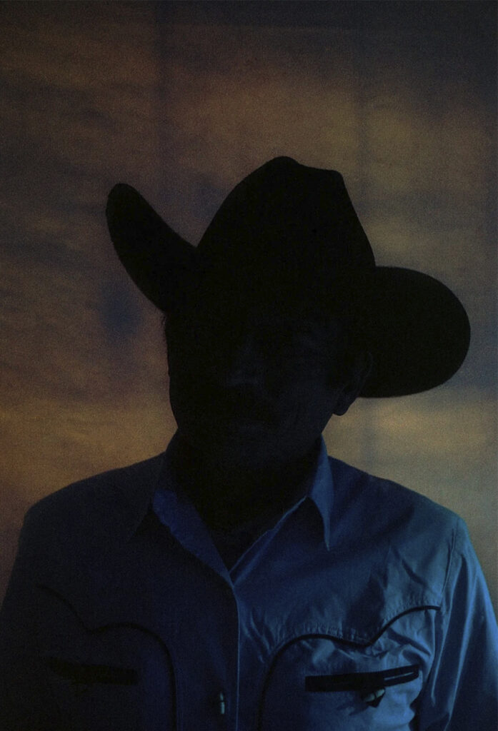 Retrato de una persona con sombrero de ala ancha y camisa clara, iluminada tenuemente sobre un fondo oscuro.