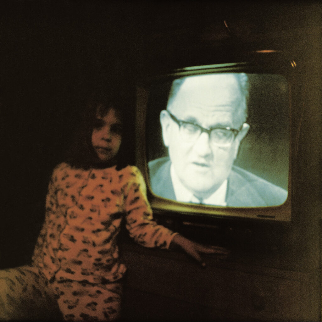 Una niña sentada junto a un televisor antiguo en una habitación oscura; en la pantalla aparece el rostro de un hombre con lentes.