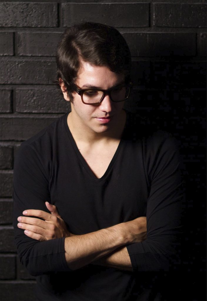 Retrato de Jakob Gómez con lentes y ropa negra frente a un muro oscuro de ladrillo.