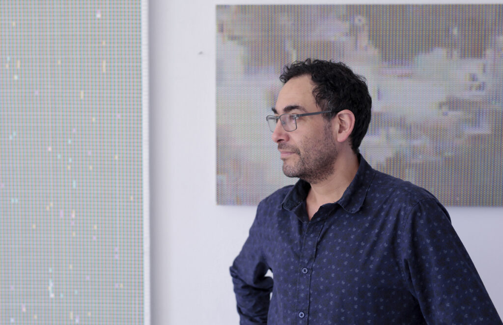 A profile portrait of Gonzalo Reyes-Araos in an exhibition space, in front of works composed of grids and RGB color patterns.