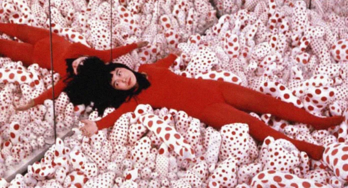 Yayoi Kusama recostada entre formas blandas cubiertas de lunares rojos y blancos, una imagen vinculada con su exploración de la repetición, la obsesión y el cuerpo.