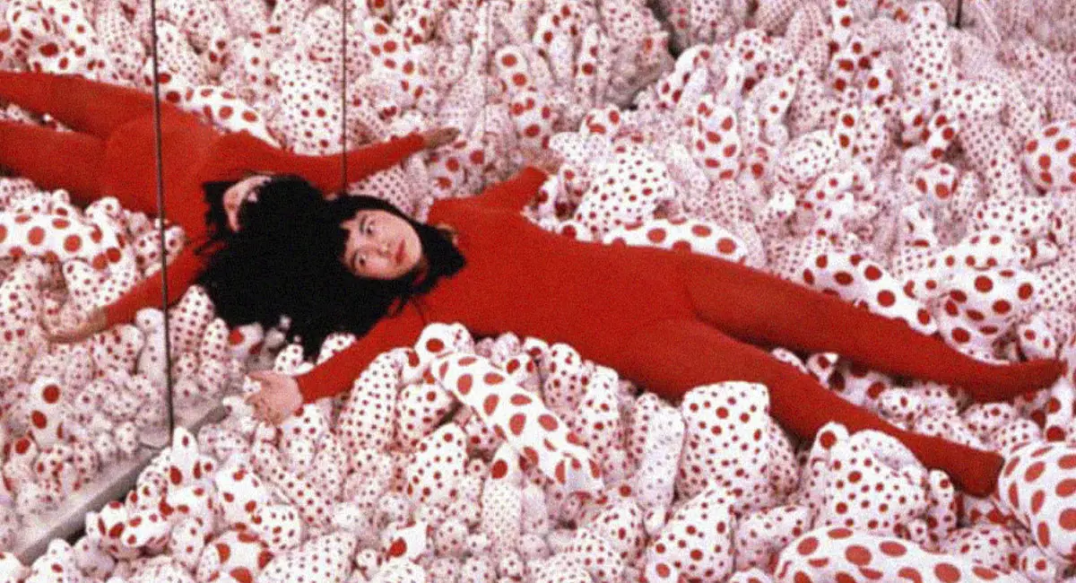 Yayoi Kusama recostada entre formas blandas cubiertas de lunares rojos y blancos, una imagen vinculada con su exploración de la repetición, la obsesión y el cuerpo.