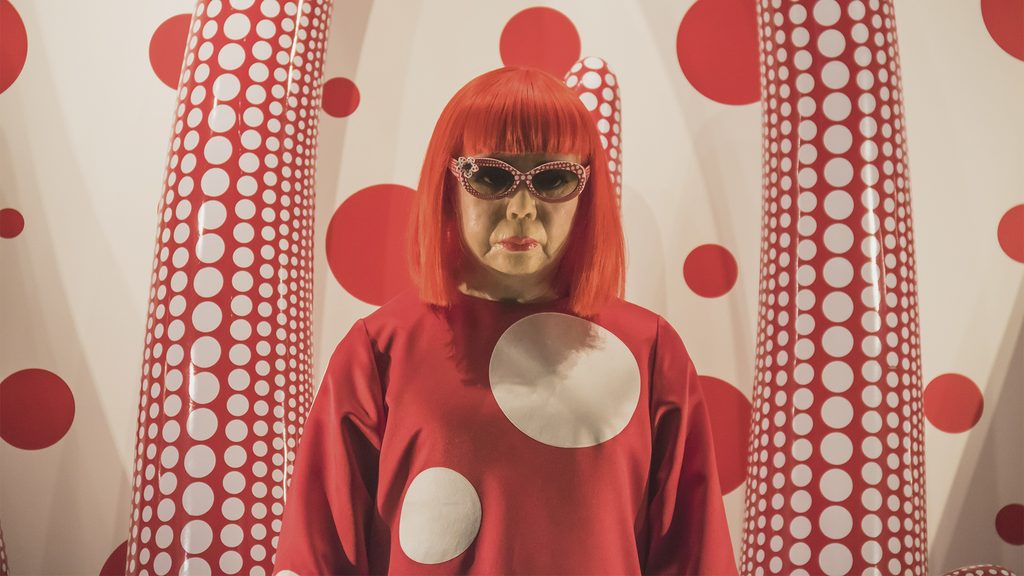 Retrato de Yayoi Kusama con peluca roja, gafas de lunares y vestido rojo, rodeada de grandes formas blancas y rojas cubiertas de puntos.