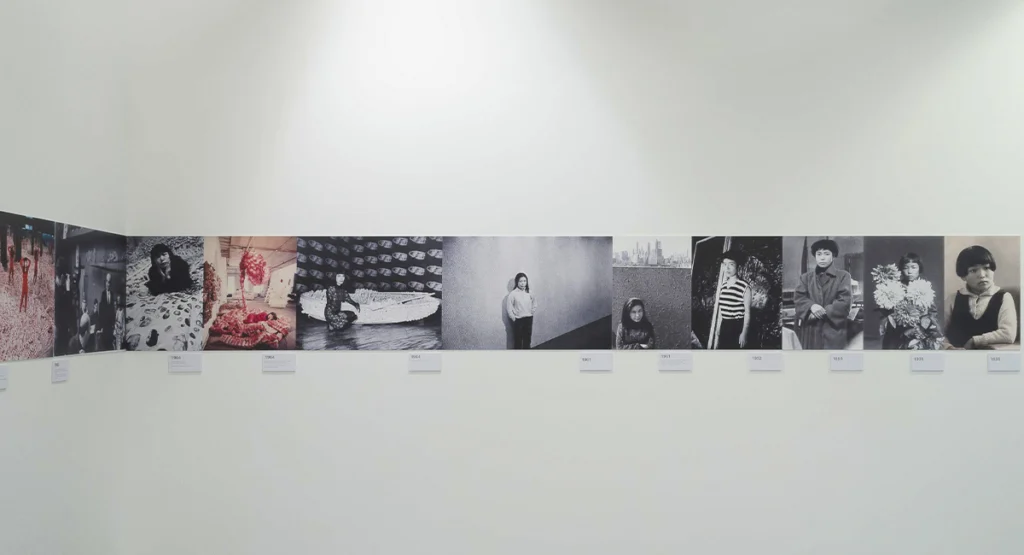 Vista de una exposición con una línea cronológica formada por fotografías de distintas etapas de la vida y carrera de Yayoi Kusama.