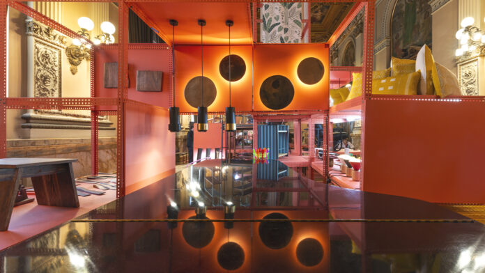 Instalación con estructuras anaranjadas, tres lámparas colgantes y muebles en un salón ornamentado.