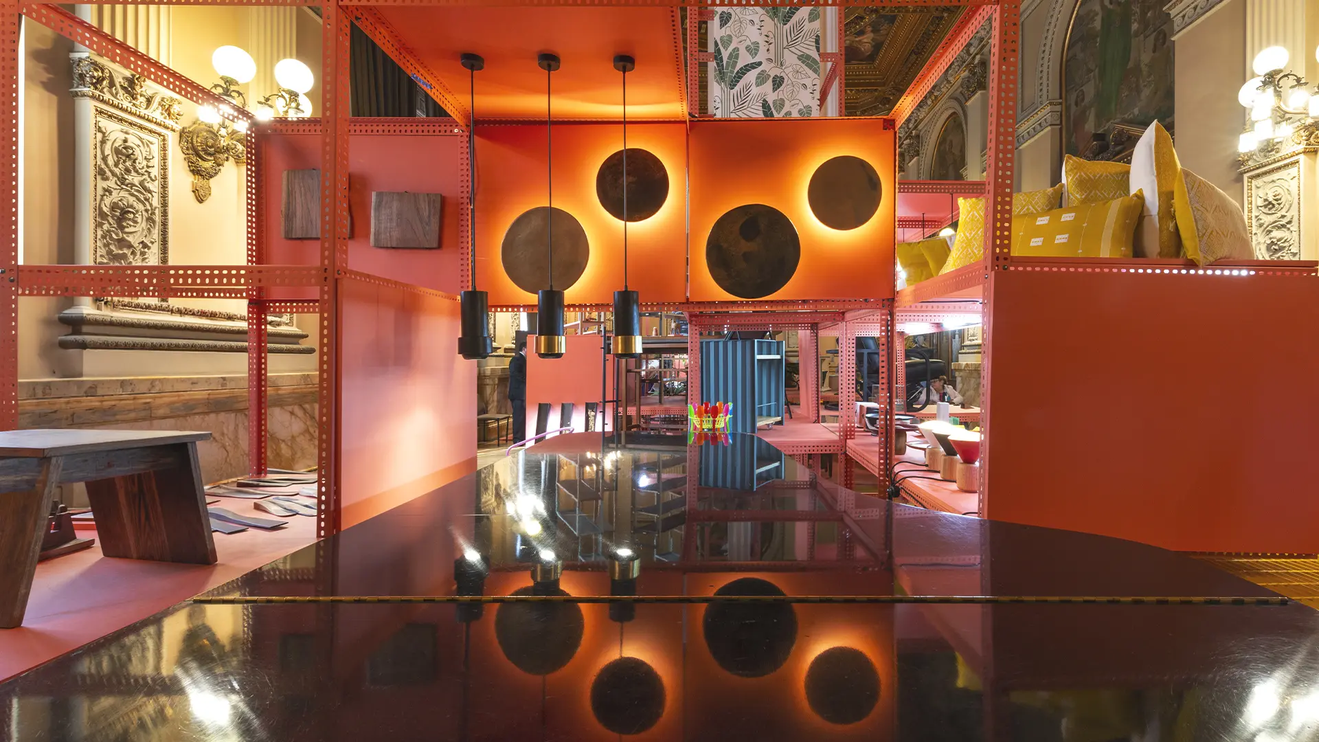 Instalación con estructuras anaranjadas, tres lámparas colgantes y muebles en un salón ornamentado.