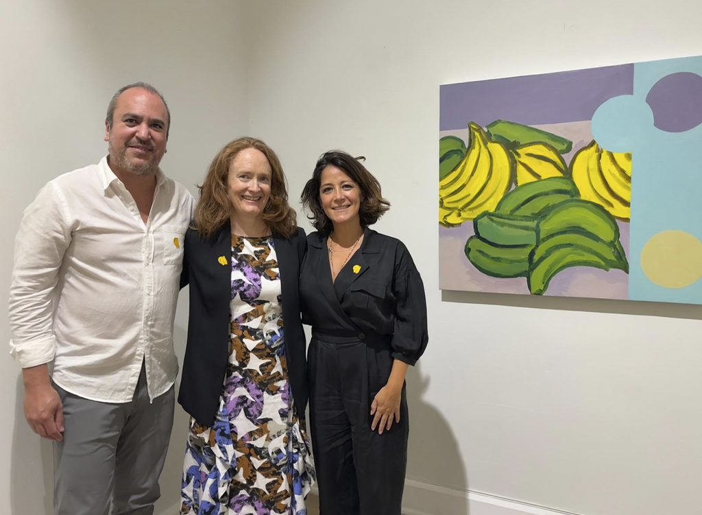 Rodrigo Borrás, Emi Winter y Andrea Walther posan frente a una pintura de plátanos de la exposición Trópico de Jersey en Galería Ethra.