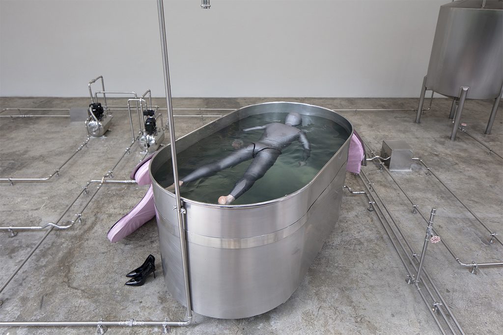 Performer con traje gris flotando dentro de una bañera metálica, rodeada por tuberías y equipo hidráulico en la galería LABOR.