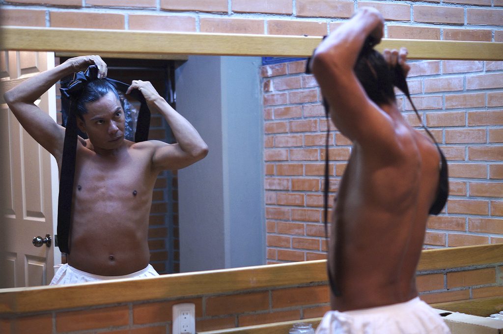Lukas Avendaño, con el torso descubierto y una falda blanca, acomoda cintas negras en su cabello frente a un espejo.
