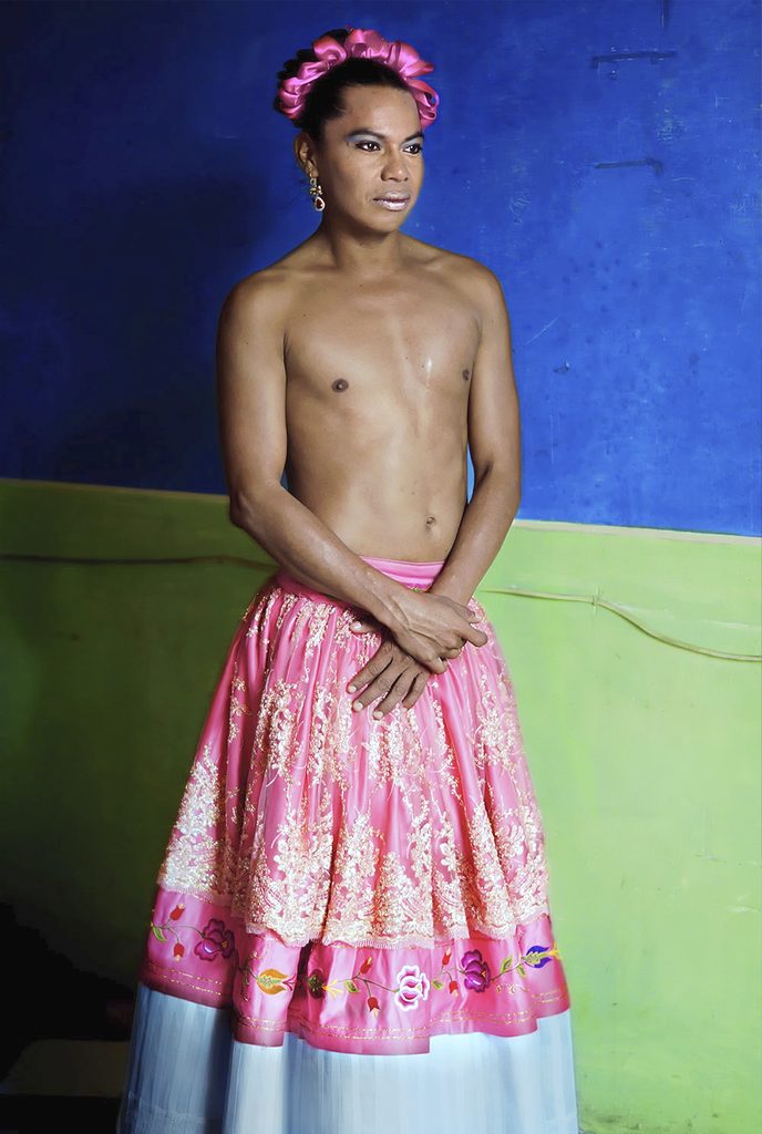 Lukas Avendaño con falda rosa bordada, cintas del mismo color en el cabello y torso descubierto, ante una pared azul y verde.