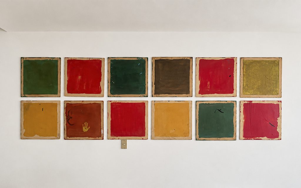 Doce cuadros cuadrados idénticos organizados en dos filas horizontales de seis en una pared blanca. Cada cuadro tiene bordes de papel y está pintado con colores sólidos como verde, rojo, amarillo, marrón y ocre.