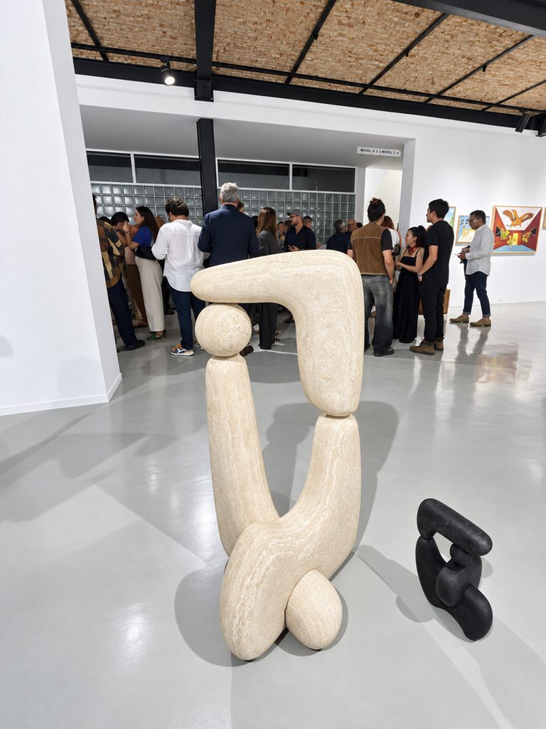 Dos esculturas orgánicas entrelazadas —una grande en cantera clara y otra pequeña en piedra oscura— colocadas directamente sobre el piso del recinto, con el público de la feria conversando al fondo