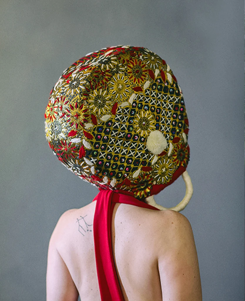 Autorretrato de Azz Rodríguez de espaldas, con la cabeza cubierta por un objeto circular bordado con motivos florales y una tela roja alrededor del cuello.