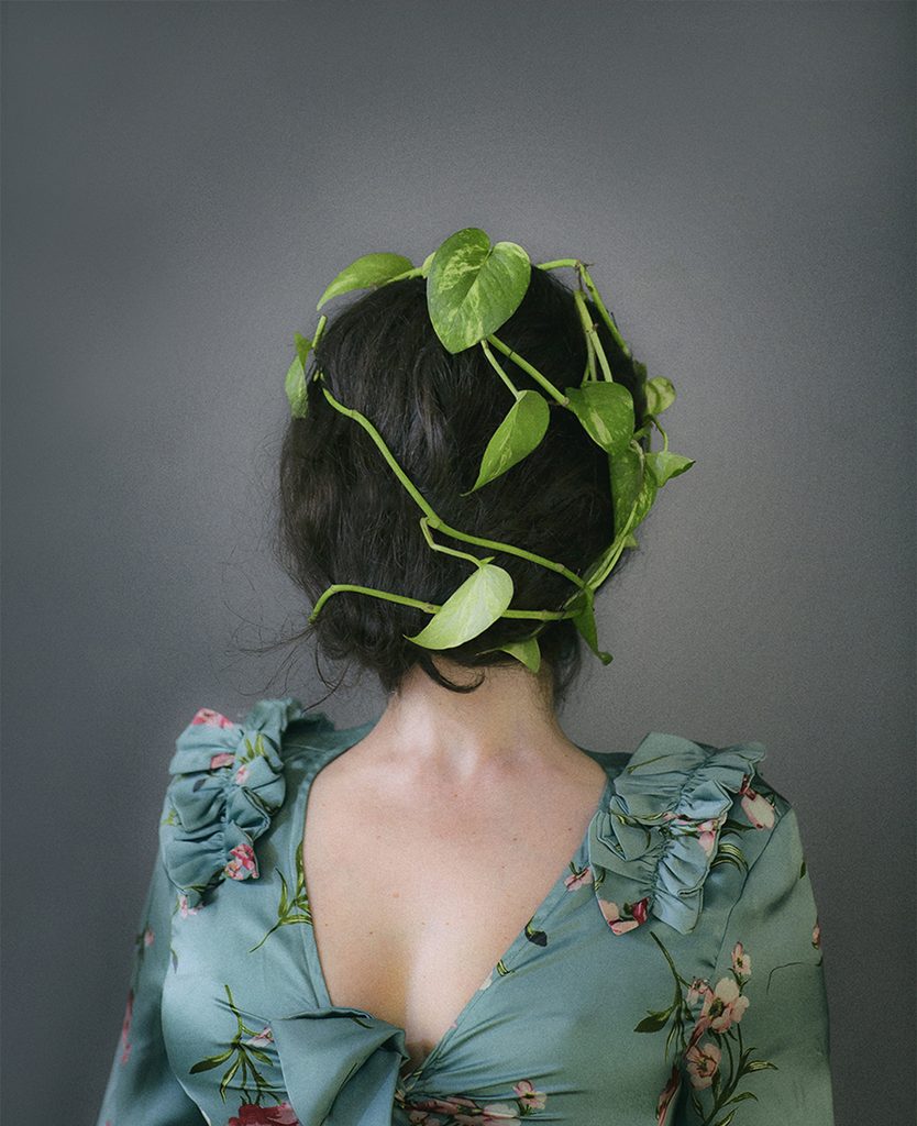 Autorretrato de Azz Rodríguez con el rostro completamente cubierto por su cabello y una planta de hojas verdes, frente a un fondo gris.