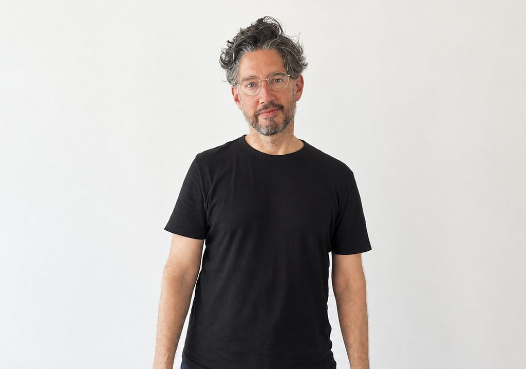 Retrato de Brett W. Schultz fundador de feria Material posando con camiseta negra y lentes sobre fondo blanco.