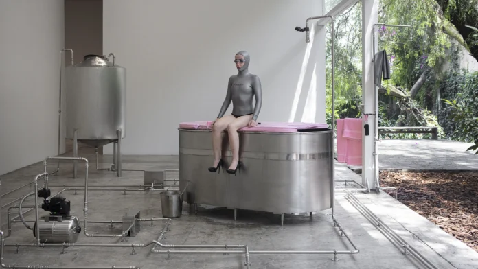Performer con traje gris, lentes y zapatos negros sentada sobre una bañera metálica conectada a un circuito hidráulico.