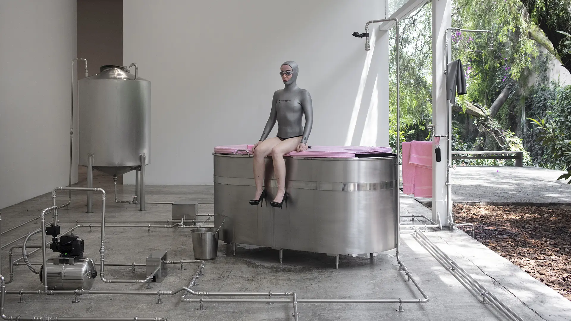 Performer con traje gris, lentes y zapatos negros sentada sobre una bañera metálica conectada a un circuito hidráulico.