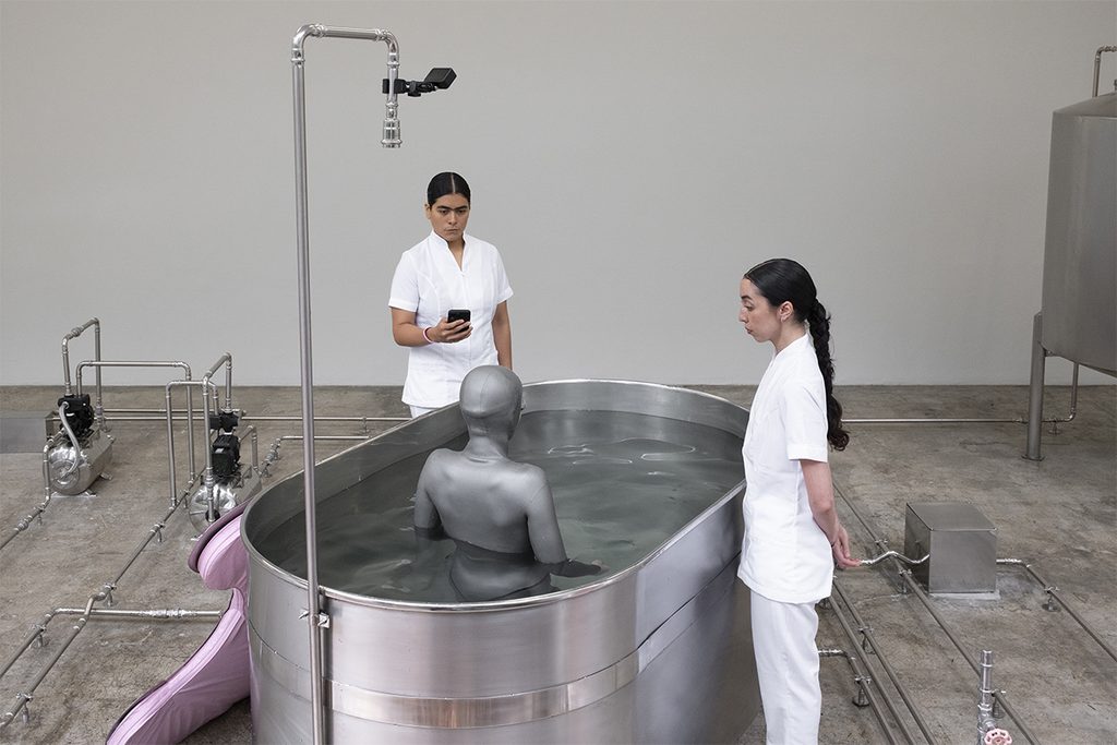 Performer con traje gris dentro de una bañera metálica, acompañada por dos asistentes vestidas de blanco.
