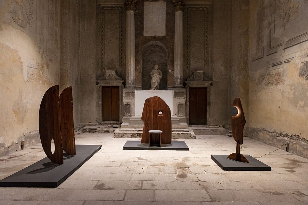 Tres piezas escultóricas de madera exhibidas en un recinto de muros antiguos.