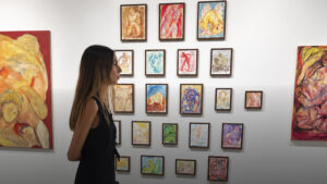 Mujer de perfil observando una colección de pequeñas pinturas coloridas colgadas en retícula en pared blanca
