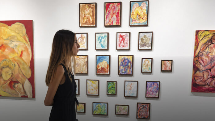 Mujer de perfil observando una colección de pequeñas pinturas coloridas colgadas en retícula en pared blanca