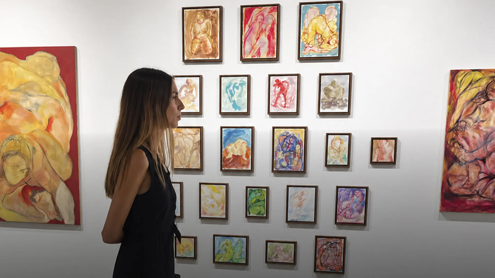 Mujer de perfil observando una colección de pequeñas pinturas coloridas colgadas en retícula en pared blanca