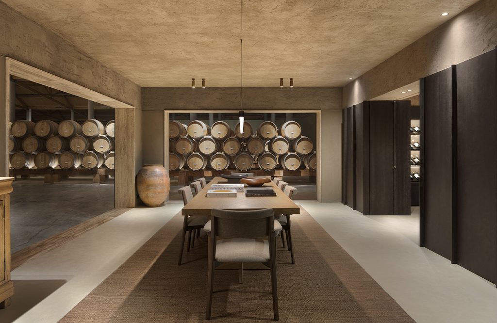 A contemporary tasting room with a central table and wine barrels stored in the back.