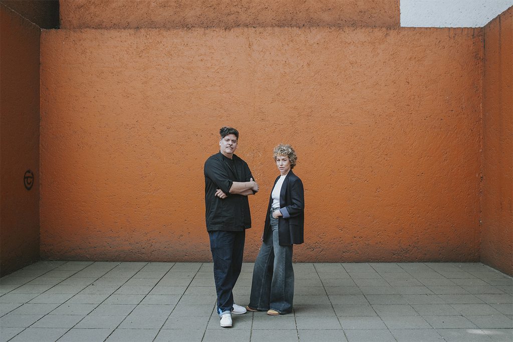 Ricardo Lozano y Joanna Ruiz Galindo posan frente a un muro anaranjado.