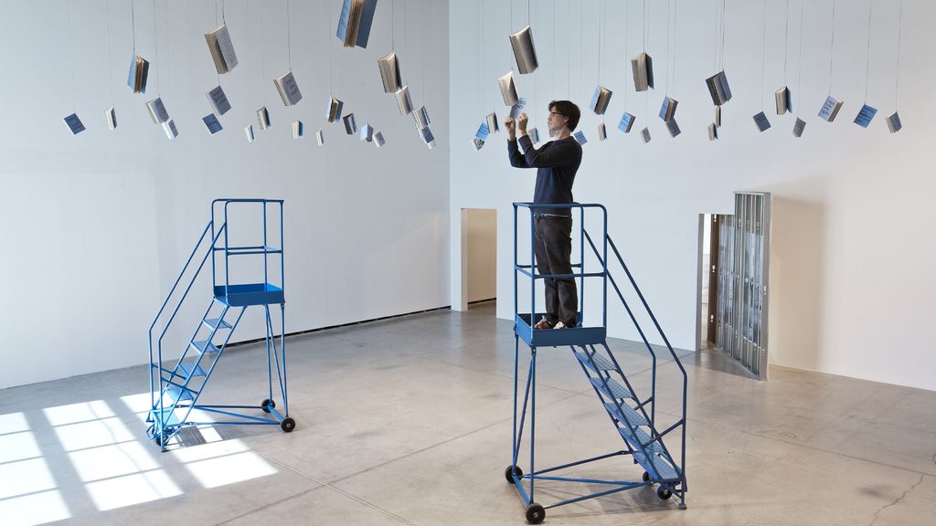 Persona sobre una escalera de plataforma ajustando una instalación de objetos suspendidos en una sala de exposición.