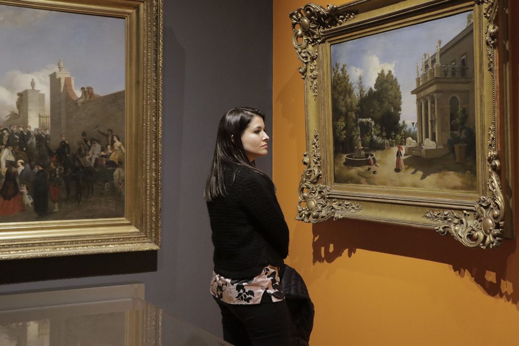 Una visitante observa una pintura de un edificio rodeado de jardines en una sala donde se exhiben cuadros con marcos dorados.