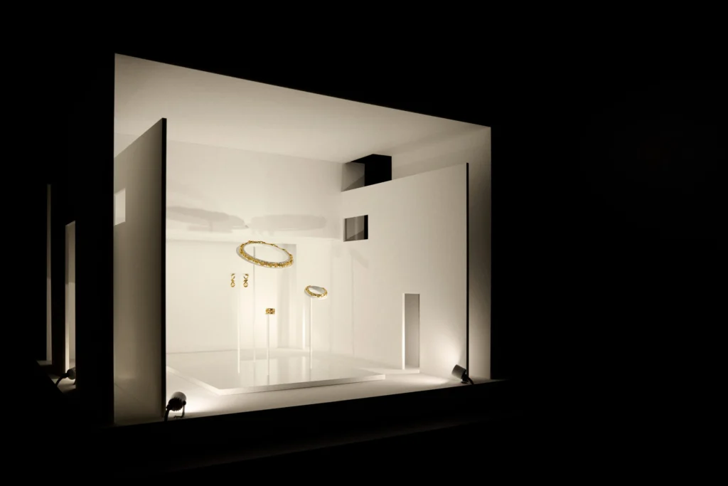 Instalación blanca con joyas doradas expuestas sobre soportes individuales e iluminación dirigida.