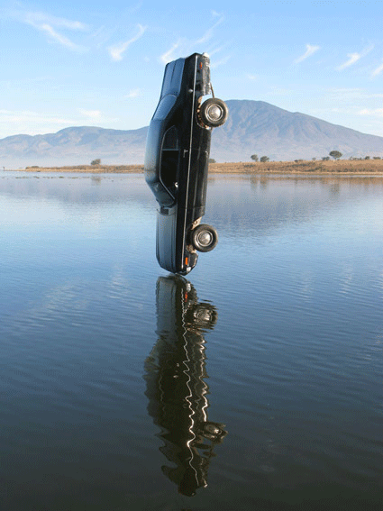 A car standing upright on a body of water, with its reflection below and mountains in the background.